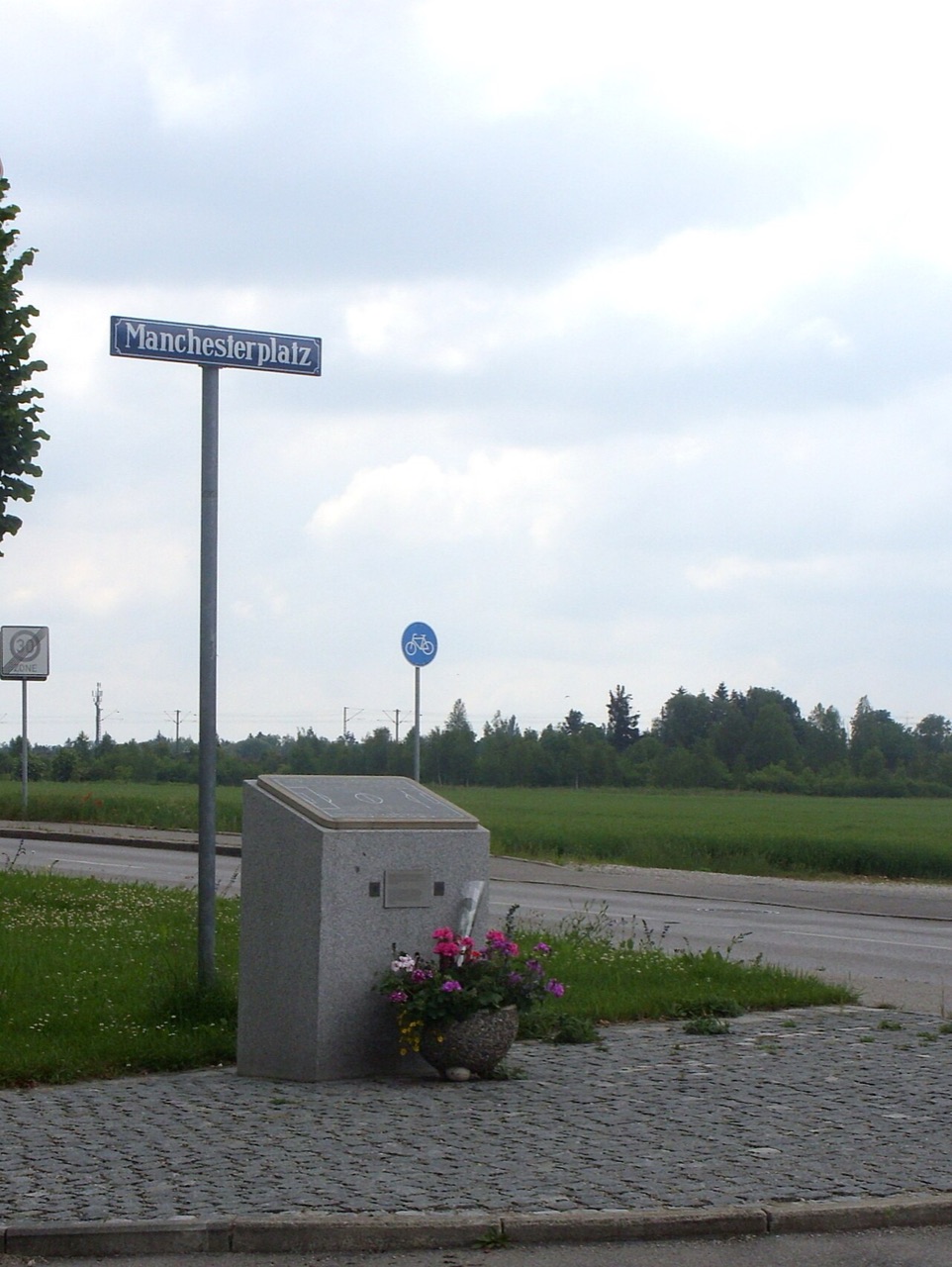 A street sign reading Manchesterplatz beside a small granite memorial with flowers